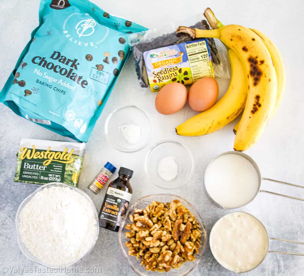Ingredients for banana bread, including bananas, eggs, dark chocolate chips, raisins, butter, flour, walnuts, baking soda, vanilla extract, milk, and a white liquid in a measuring cup.