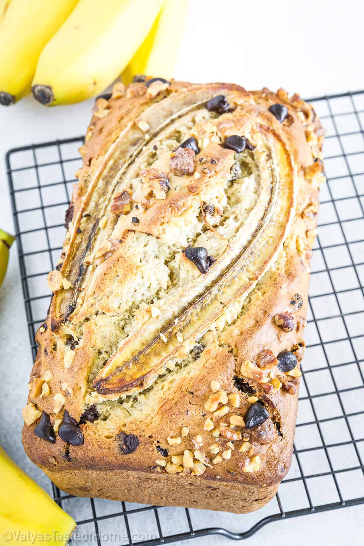 Loaf of banana bread topped with sliced banana, walnuts, and chocolate chips, resting on a cooling rack with bananas nearby.