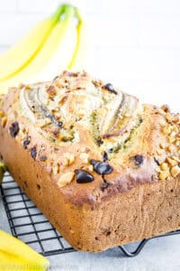 A loaf of banana bread topped with banana slices, chocolate chips, and chopped walnuts sits on a cooling rack with bananas in the background.
