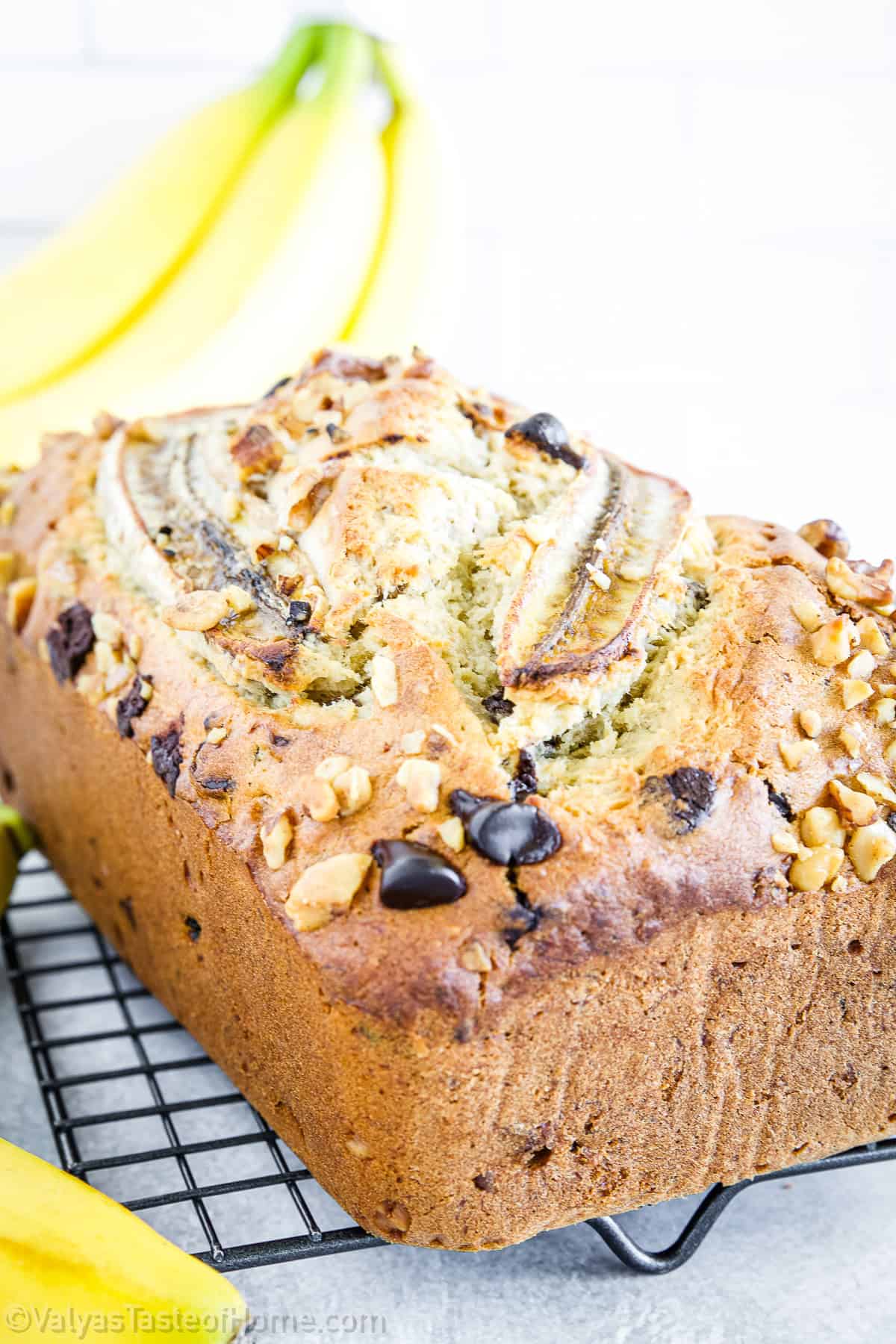 A loaf of banana bread topped with banana slices, chocolate chips, and chopped walnuts sits on a cooling rack with bananas in the background.