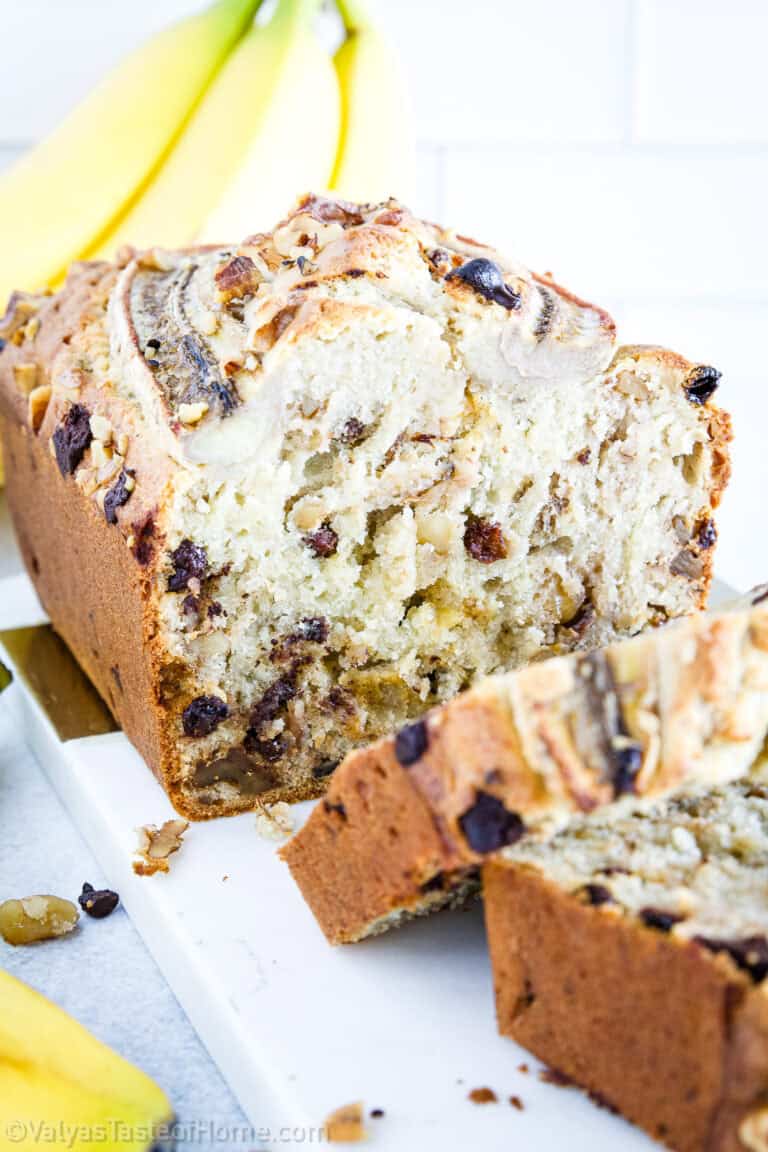 A loaf of banana nut bread with visible banana slices and nuts, partially sliced, sits on a white cutting board with bananas in the background.