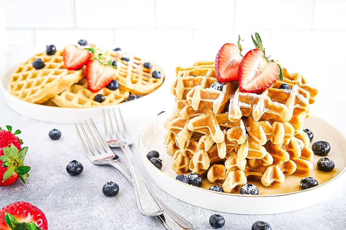 A stack of waffles topped with sliced strawberries and blueberries, served on a white plate with extra fruit and forks on the side.