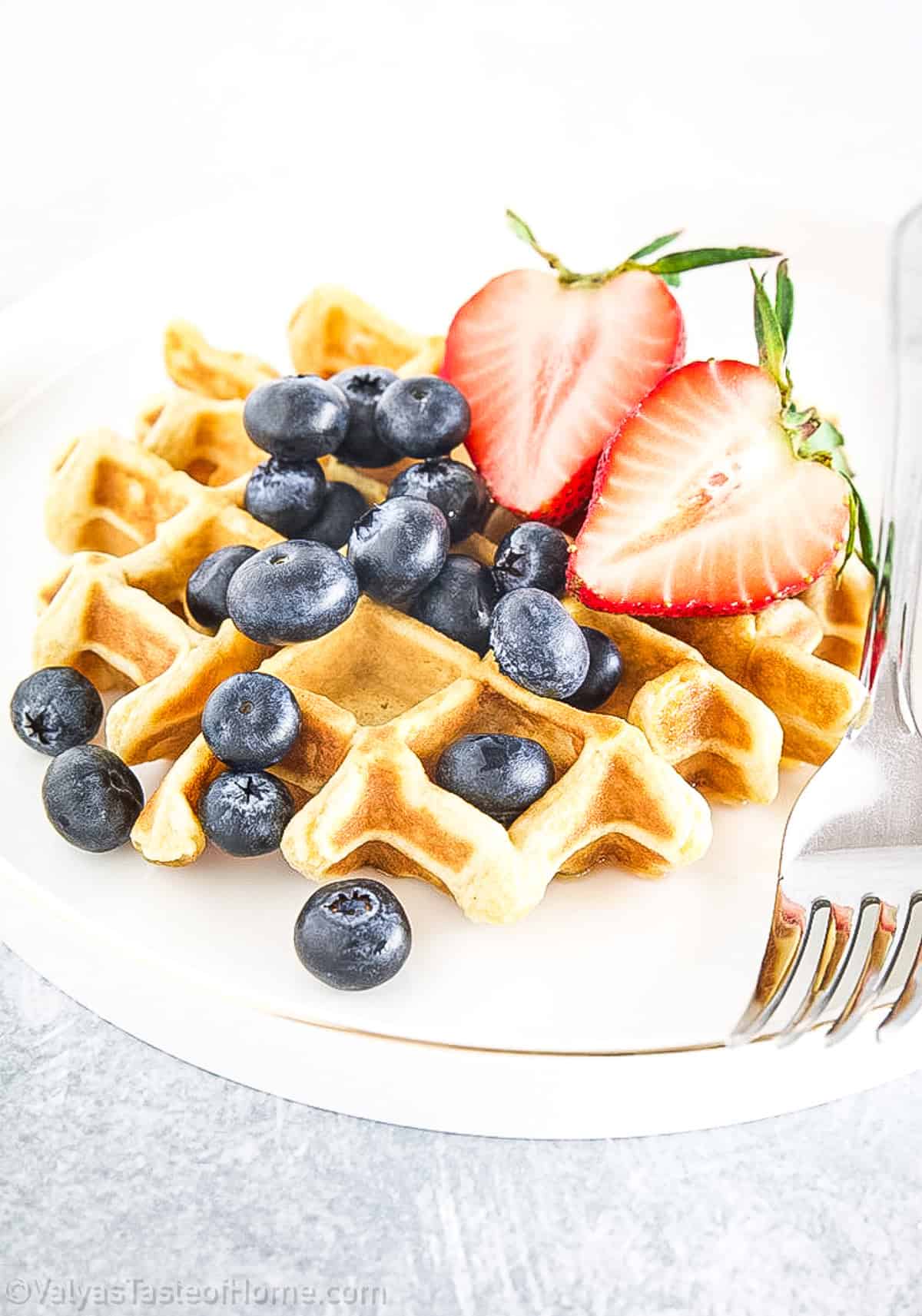A plate with a waffle topped with blueberries and two halved strawberries, with a fork on the side.