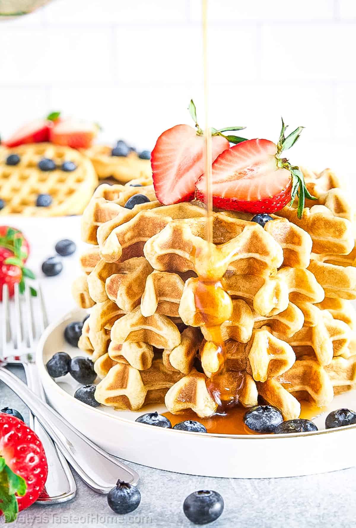 A stack of waffles topped with sliced strawberries and blueberries is being drizzled with syrup, with more fruit and waffles visible in the background.