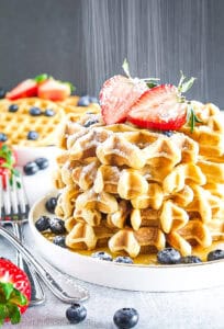 A stack of waffles topped with powdered sugar and sliced strawberries, surrounded by blueberries on a white plate.