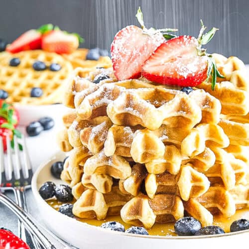 A stack of waffles topped with powdered sugar and sliced strawberries, surrounded by blueberries on a white plate.