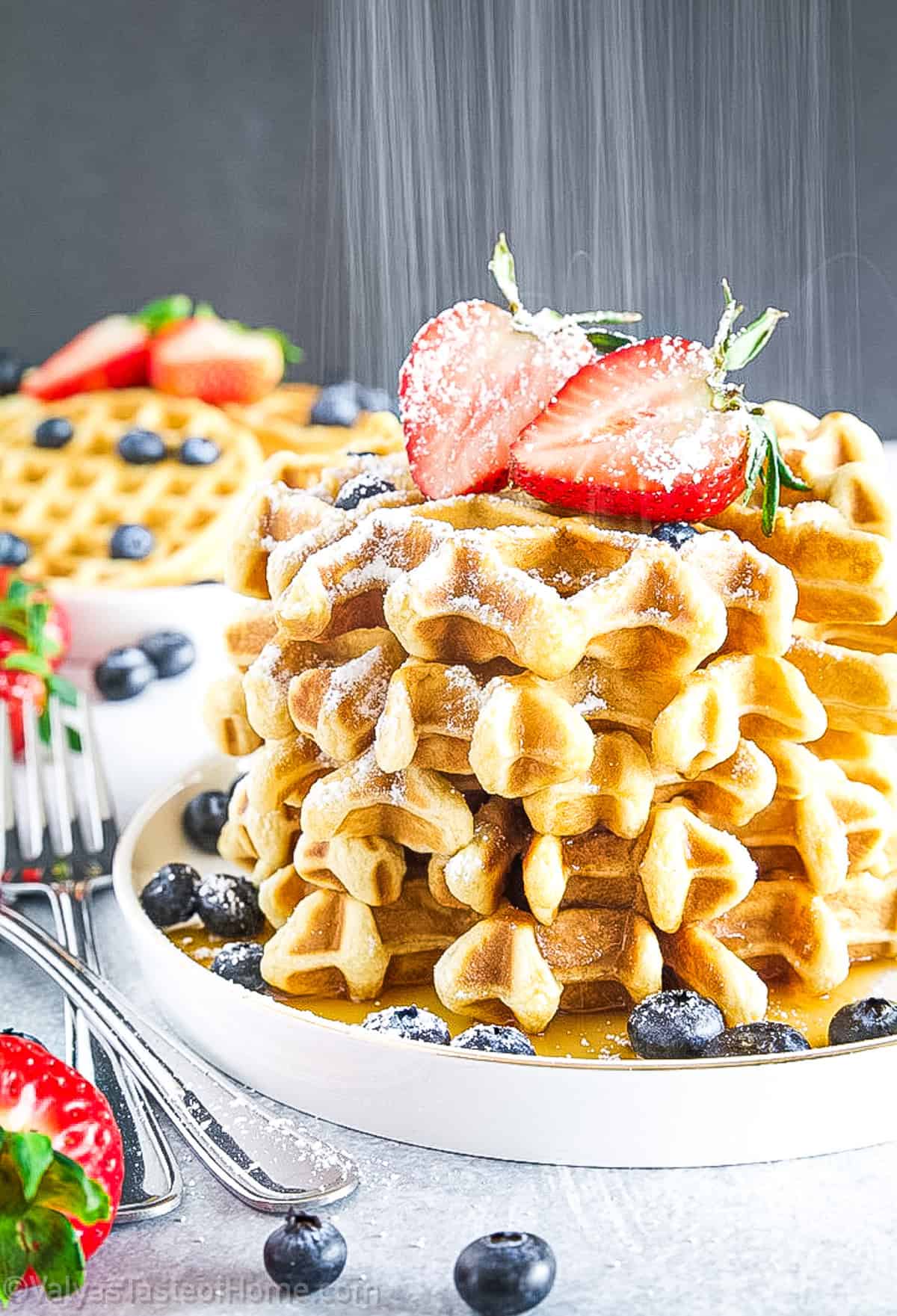 A stack of waffles topped with powdered sugar and sliced strawberries, surrounded by blueberries on a white plate.