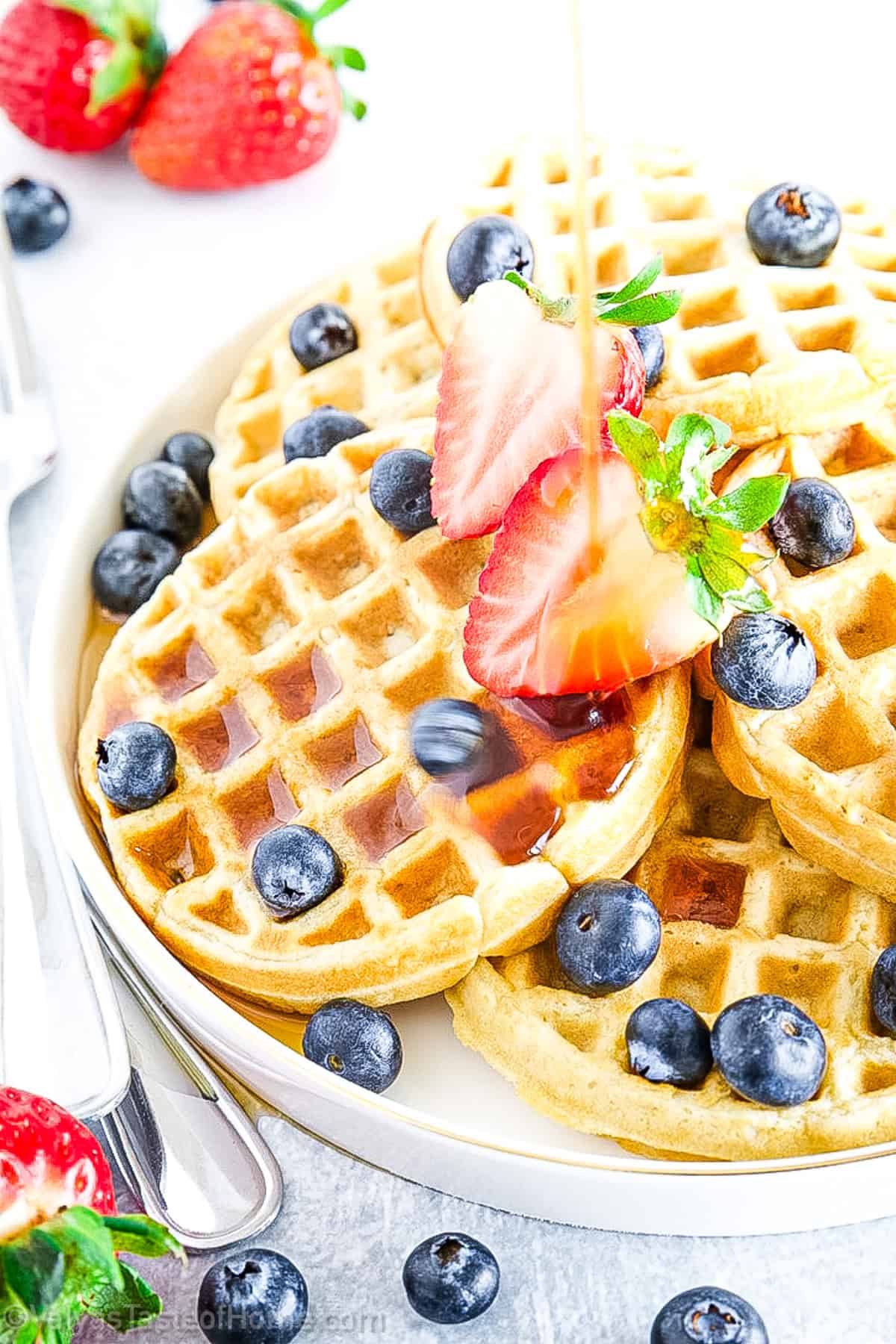 A plate of waffles topped with fresh strawberries, blueberries, and syrup being poured, with a fork and knife beside the plate.