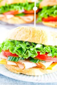 A turkey croissant sandwich with lettuce, tomato, cucumber, turkey, and cheese on a white plate, with more croissant sandwiches on a tray in the background.