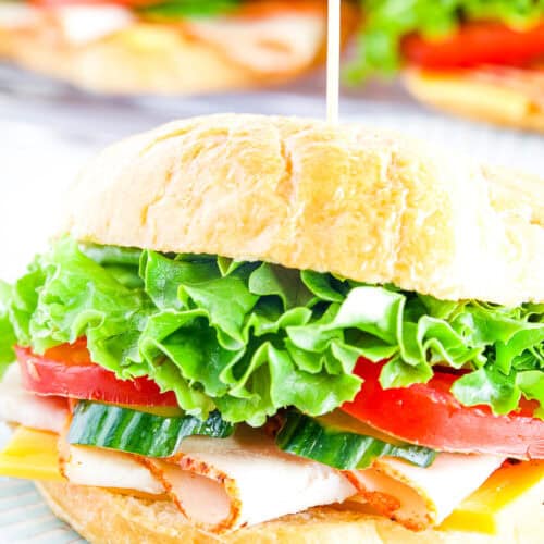 A turkey croissant sandwich with lettuce, tomato, cucumber, turkey, and cheese on a white plate, with more croissant sandwiches on a tray in the background.