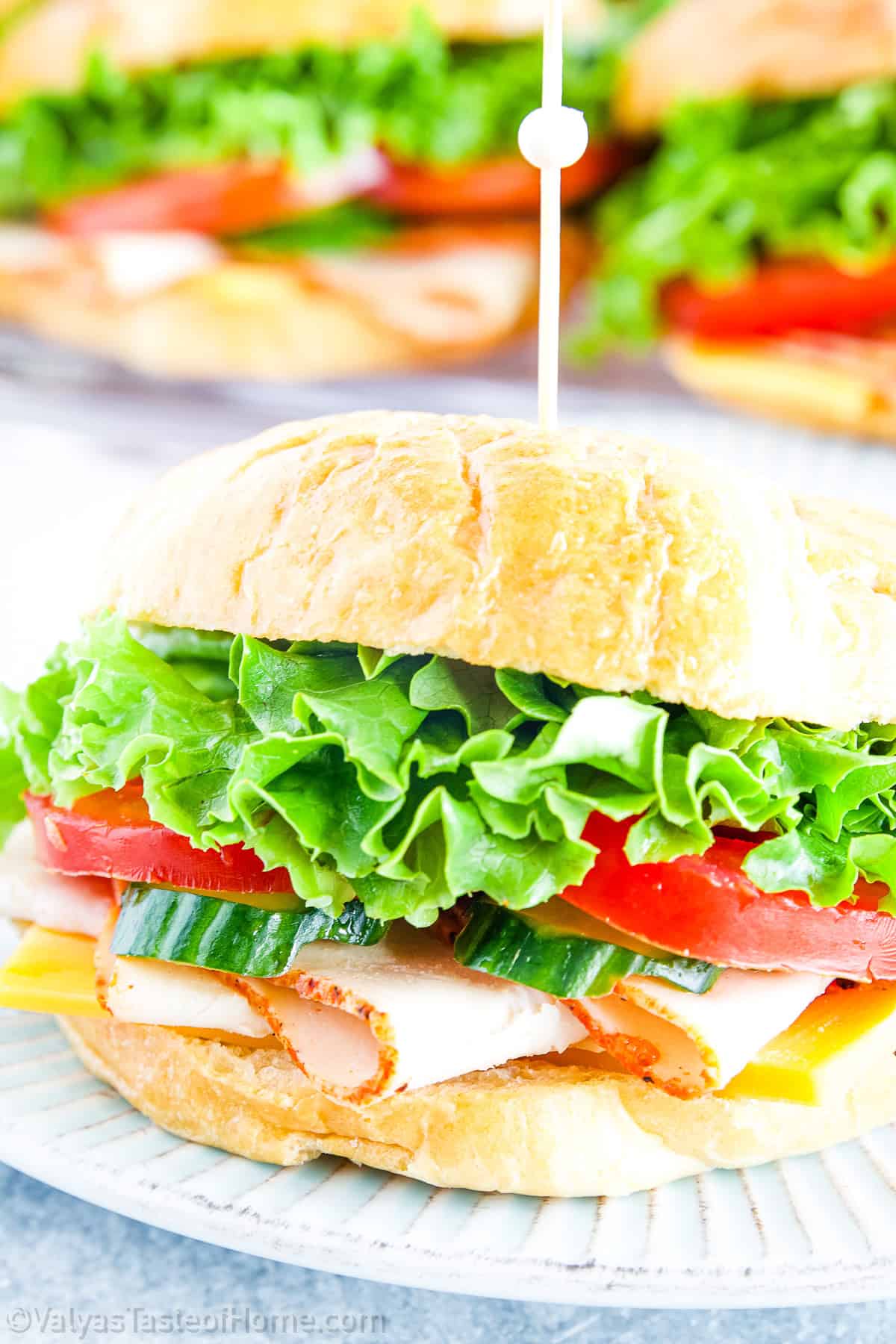 A turkey croissant sandwich with lettuce, tomato, cucumber, turkey, and cheese on a white plate, with more croissant sandwiches on a tray in the background.