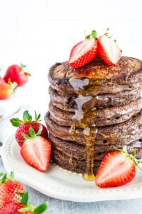 A stack of chocolate pancakes topped with halved strawberries and drizzled with syrup, served on a white plate with additional strawberries.