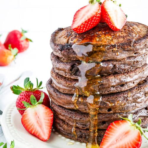 A stack of chocolate pancakes topped with halved strawberries and drizzled with syrup, served on a white plate with additional strawberries.