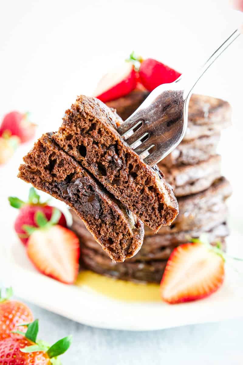A fork holds a stack of sliced sourdough discard chocolate pancakes in front of a pile of chocolate pancakes with strawberries on a plate.