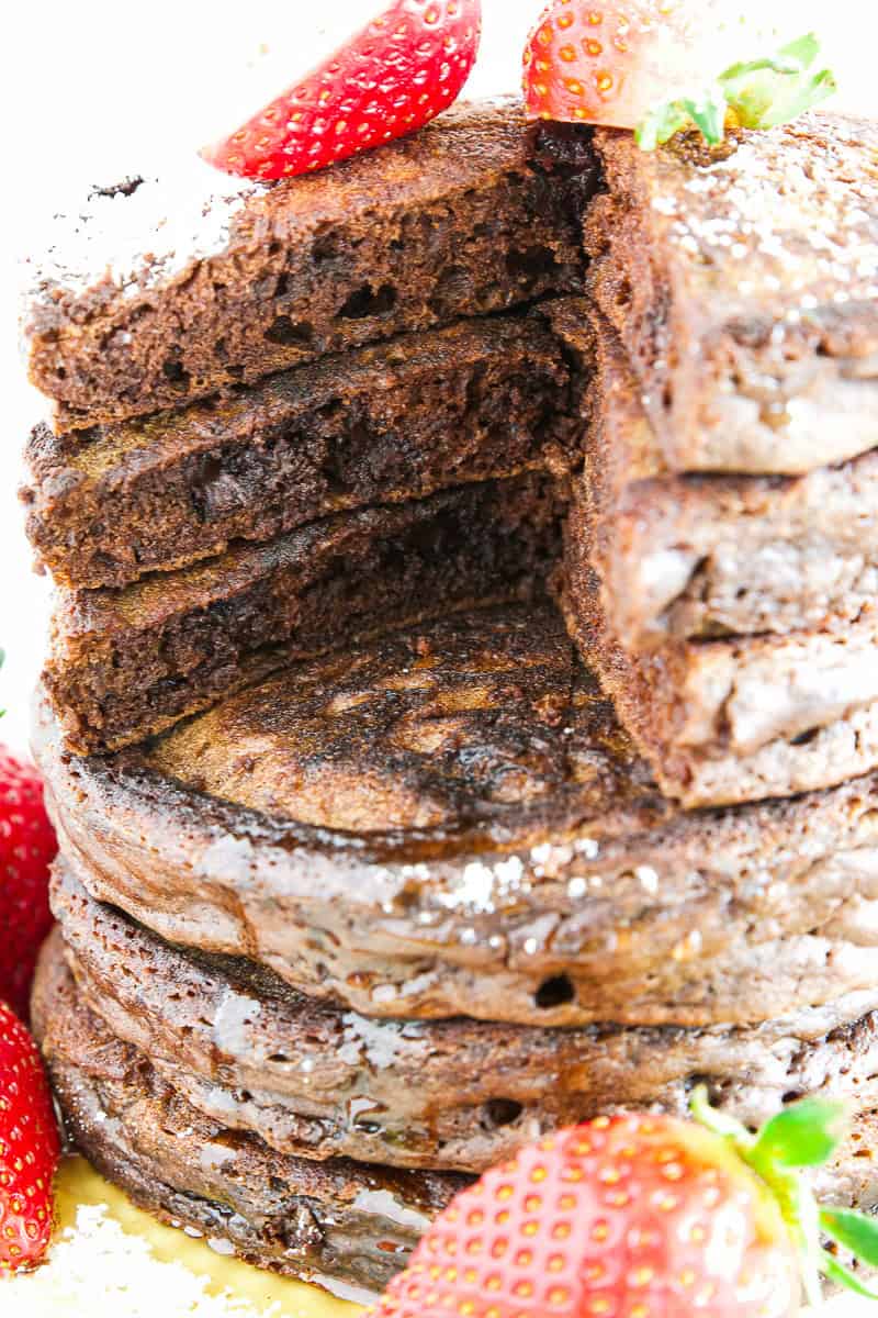 A stack of thick chocolate pancakes with a wedge cut out, topped and surrounded by fresh strawberries, dusted with powdered sugar.